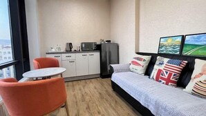 Un espacio habitable compacto con kitchenette, un sofá con cojines decorativos, una pequeña mesa redonda y una sola silla naranja.