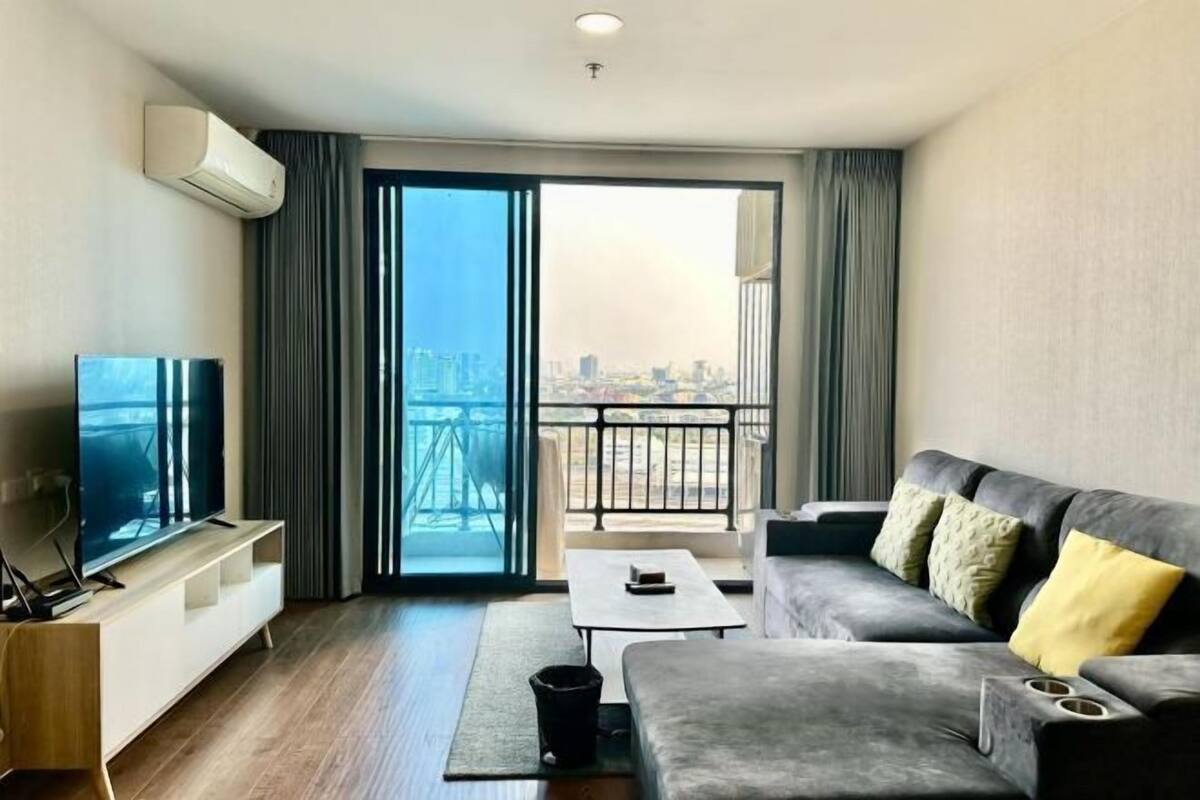 A modern living room with a flat-screen TV, a sofa with cushions, a coffee table, and a view of a cityscape through a large sliding glass door.