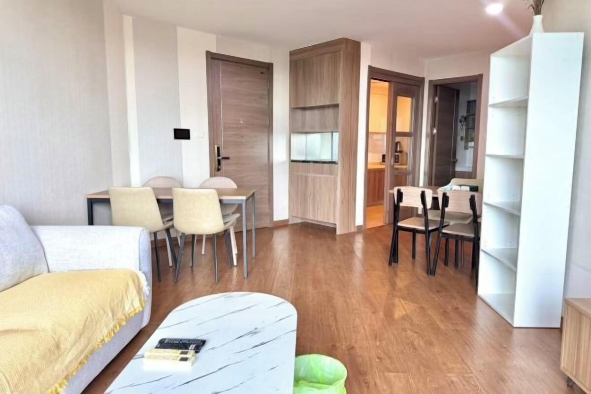 A compact living space with a bed, a dining table, and chairs. There is a small seating area with a sofa and a coffee table. The room has wooden flooring and a bookshelf.