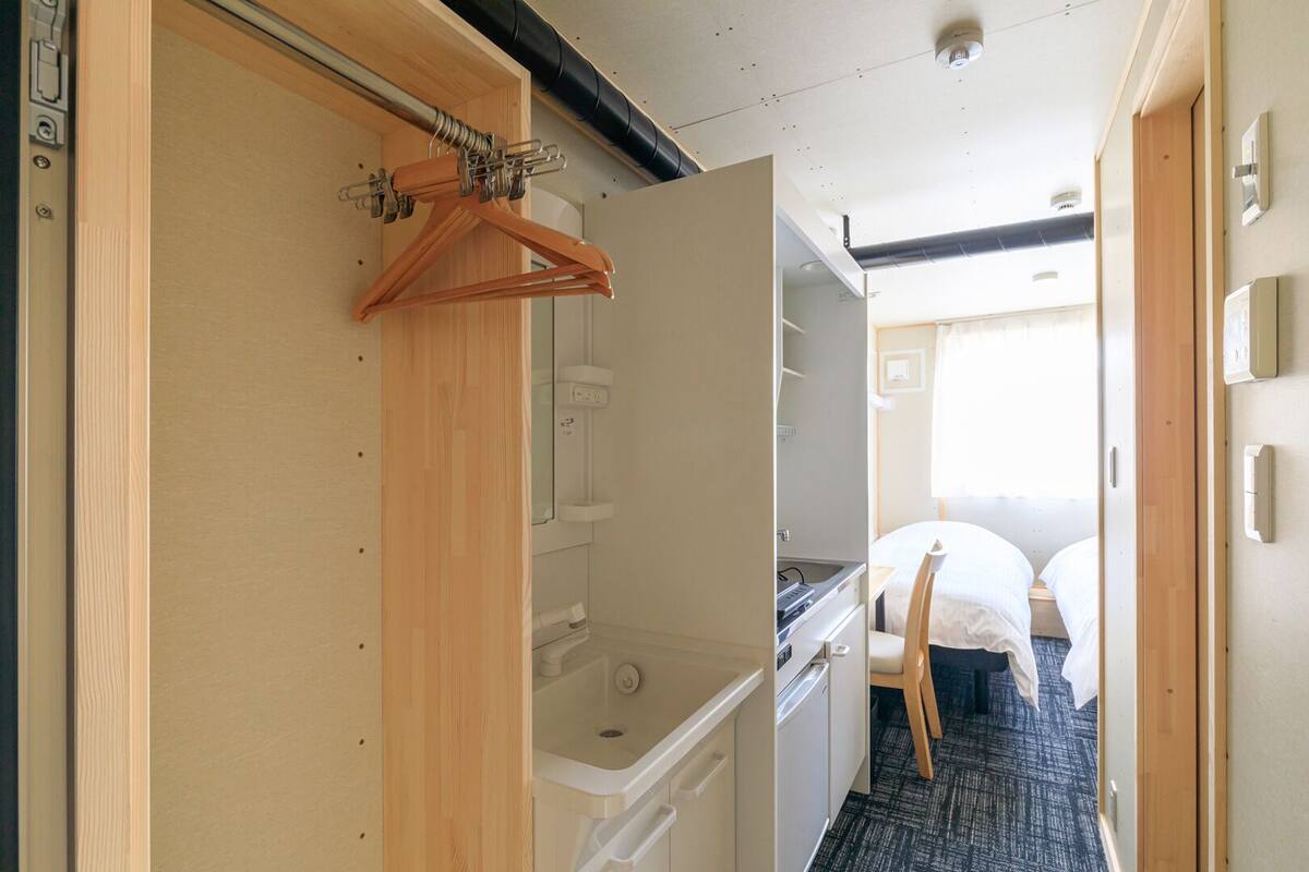 Un interior compacto y bien iluminado, con un armario de madera, una pequeña cocina y una cama con cabecera.