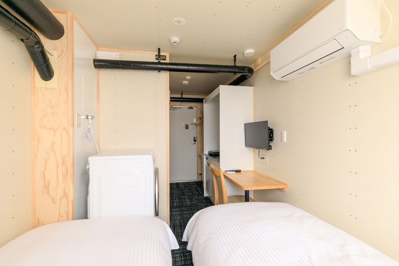 Habitación de hotel compacta con dos camas individuales, una kitchenette pequeña, un televisor montado en la pared y un equipo de aire acondicionado instalado en el techo.
