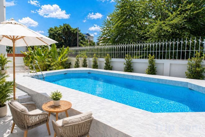 A swimming pool with a white tiled edge, surrounded by a white fence, and a small table with chairs and a potted plant.