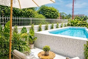 Une piscine dont le bord est recouvert de carreaux blancs, entourée de plantes en pot et de mobilier d’extérieur.
