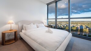 A modern hotel room with a large bed, a nightstand, a lamp, and a view of a cityscape through large windows.