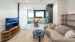 A modern living room with a flat-screen TV on a wooden stand, a round coffee table, a beige sofa, and a balcony with a city view.