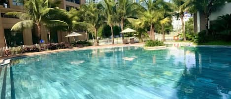 Une piscine entourée de palmiers et délimitée par une clôture, avec des immeubles de grande hauteur en arrière-plan.