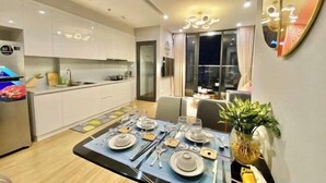 A modern dining area with a table set for four, a kitchen in the background, and a large window with curtains. - Vinhomes Skylake Premier Residence Ha Noi - Marina Housing (Hanoi)