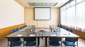 Ett konferensrum med ett långt bord, blå stolar, en projektorduk och ett stort fönster med gardiner.