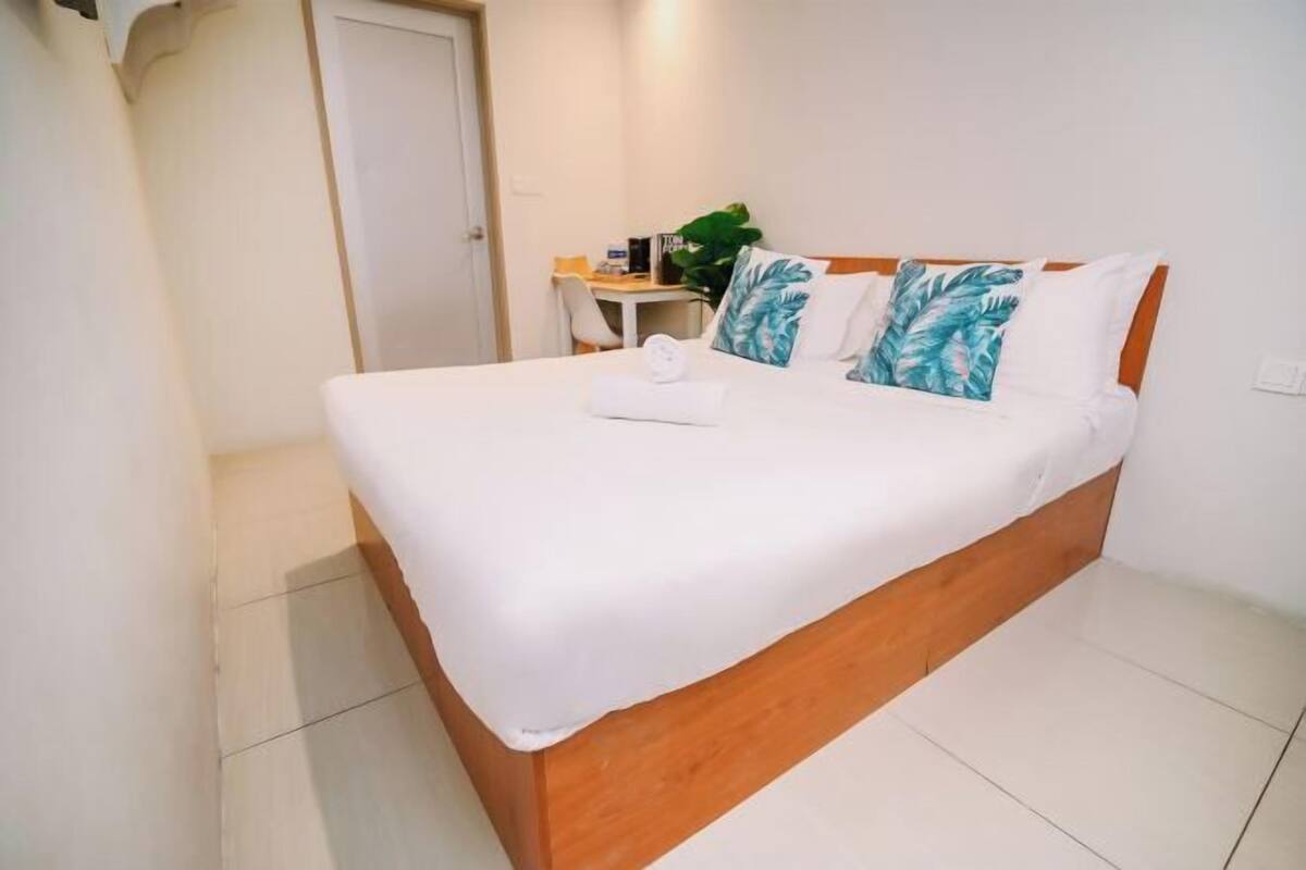 Ein ordentlich bezogenes Bett mit Holz-Kopfstück, ein kleiner Schreibtisch mit Stuhl und ein Blumentopf im Hotelzimmer.