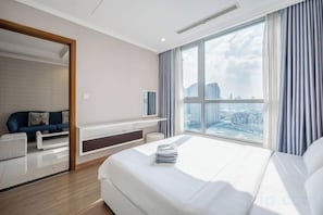 A modern hotel room with a large bed, a seating area with a sofa, a TV, and a view of the cityscape. - The Landmark Collection Suite (Ho Chi Minh City)