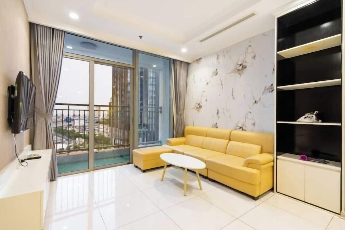 A modern living room with a yellow sofa, a white coffee table, a TV on a stand, and a large sliding glass door leading to a balcony.