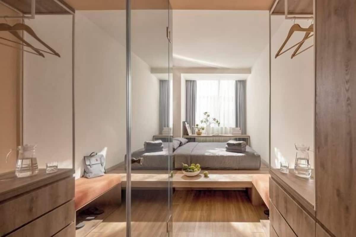Ein modernes Schlafzimmer mit Glastrennwand, einem Bett mit grauer Bettwäsche, einem Nachttisch mit Lampe und einem Fenster mit Vorhängen.