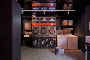 A laundry facility with multiple washing machines and dryers, organized shelves with baskets, and a small seating area. - Express by M Village Global City, a brand of Modern Village Lifestyle (Ho Chi Minh City)