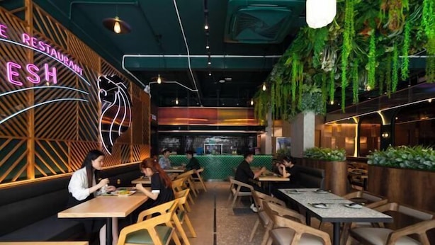 Interior de restaurante con mesas y sillas de madera, un letrero de neón y plantas colgantes.