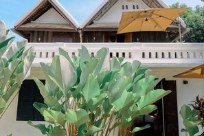 Un bâtiment traditionnel doté d’un balcon, avec de grandes plantes vertes au premier plan et un parasol jaune.