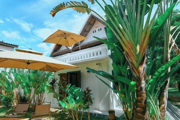 A white building with a tiled roof, surrounded by lush greenery and outdoor umbrellas.