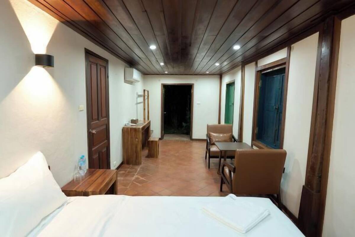 A hotel room with a bed, a small table, a chair, a wooden cabinet, and a door leading to another room.