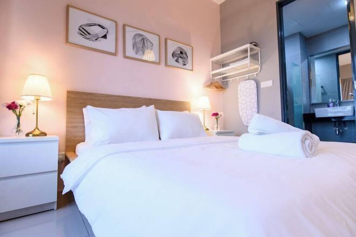 Une chambre d’hôtel avec un lit, deux tables de chevet avec des lampes, une étagère fixée au mur, un miroir et une salle de bain visible à travers une porte ouverte.