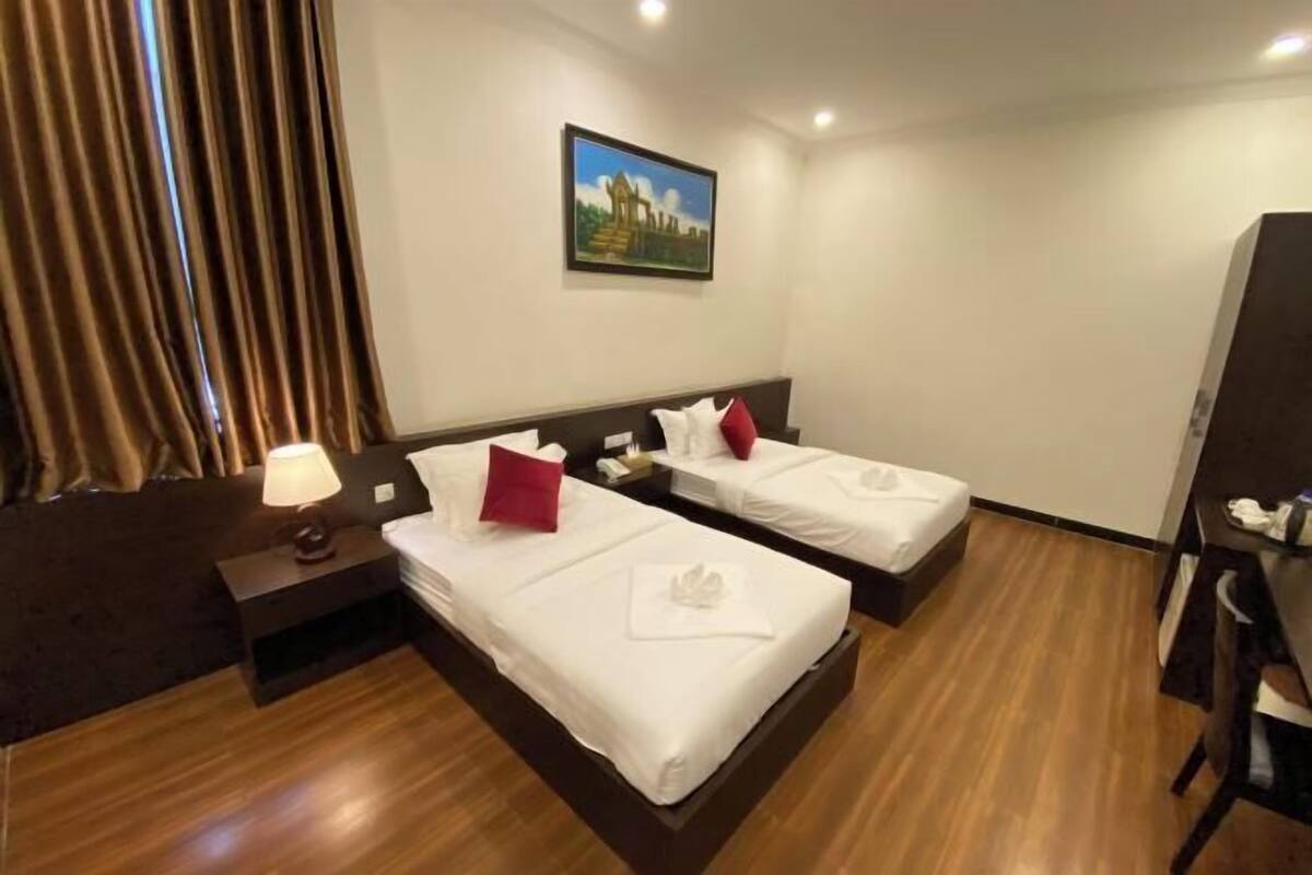A hotel room with two beds, a nightstand with a lamp, a painting on the wall, and a desk with a chair.