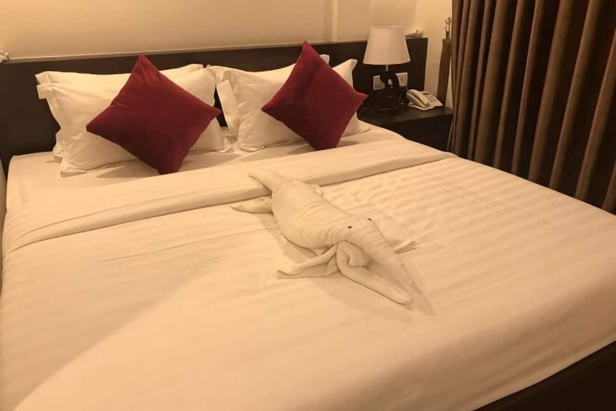 A neatly made bed with white linens and two red pillows, a white towel, and a nightstand with a lamp and phone.