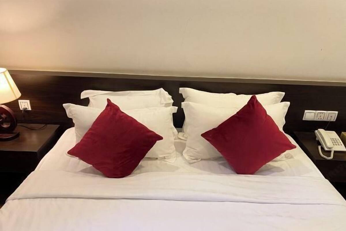 A hotel room with a neatly made bed, two red pillows, a bedside lamp, and a phone on the nightstand.