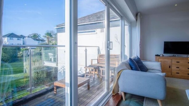 A modern living room with a large sliding glass door leading to a deck, a sofa with cushions, a television, and wooden furniture. - Footsteps - 2 Bedroom Apartment - Saundersfoot (Saundersfoot)