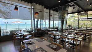Un restaurant amb taules i cadires preparades per dinar, grans finestres que donen a una vista del mar i llums penjants al sostre.