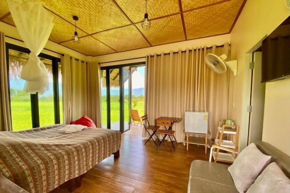 A bedroom with a bed, a sofa, a table, and a chair. There is a window with curtains and a view of the outdoors.