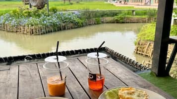 A wooden table with two drinks and a plate of food, overlooking a calm water body and a rural landscape.