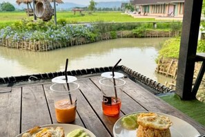 A wooden table with two drinks and a plate of food, overlooking a calm water body and a rural landscape. - Baan Porhdoi Goidao (Chiang Dao)