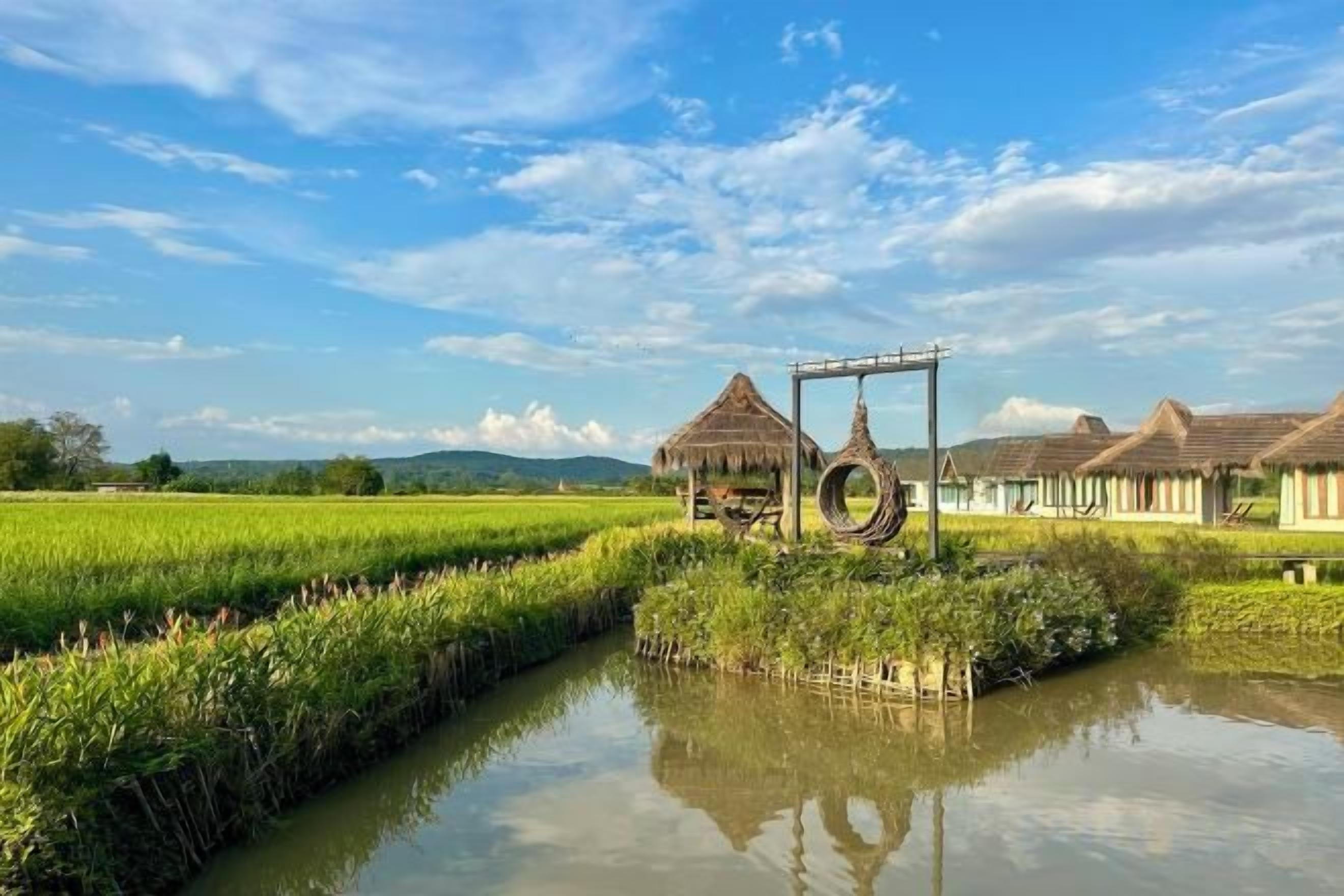 A rural landscape with a water channel, traditional thatched-roof structures, and a clear sky.