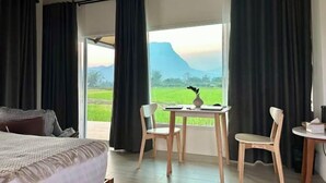 A bedroom with a bed, a table with chairs, and a view of a green field and mountains through a window. - Baan Porhdoi Goidao (Chiang Dao)