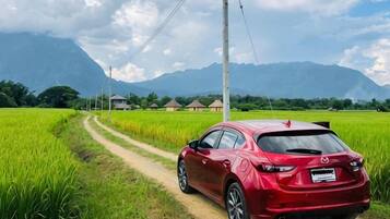 A red car parked on a rural dirt road with green fields and mountains in the background.