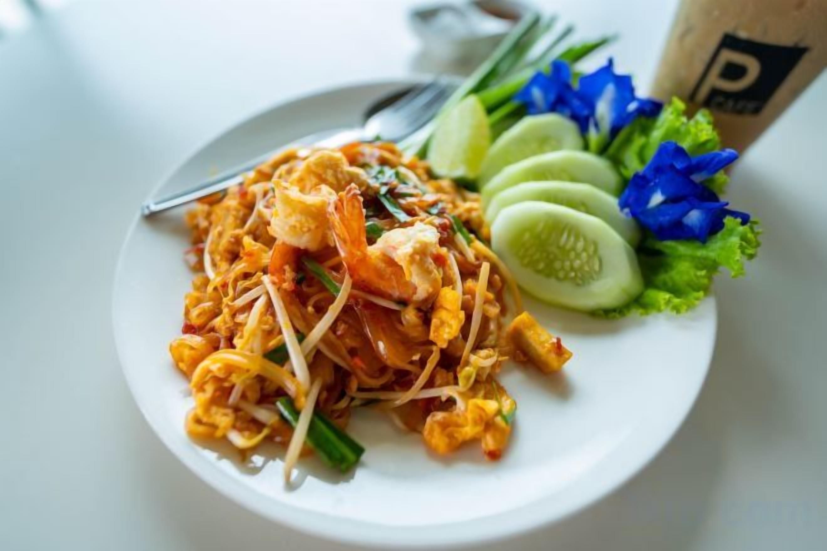 A plate of Pad Thai noodles with shrimp, bean sprouts, and a side of cucumber slices and lettuce.