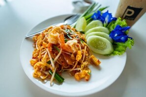 Eine Portion Pad Thai Nudeln mit Garnelen, Sojasprossen, dazu Gurkenscheiben und Salat.