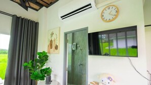A room with a wooden ceiling, a clock, a window with a view of greenery, a door, and a small table with a plant and a stuffed toy. - Baan Porhdoi Goidao (Chiang Dao)