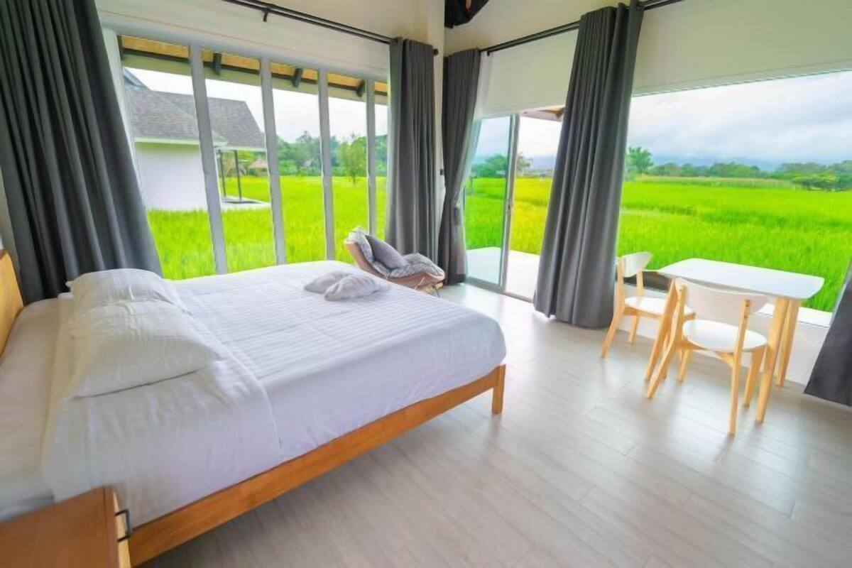 A bedroom with a bed, a table, and chairs, large windows with curtains, and a view of a green field.