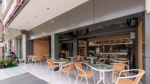 An outdoor seating area with tables and chairs, a motorcycle parked to the side, and a building entrance with glass doors. - Banana Guesthouse (Pattaya)