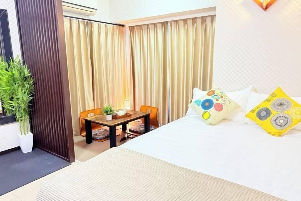 A hotel room with a bed, a small table with chairs, a potted plant, and a curtain.