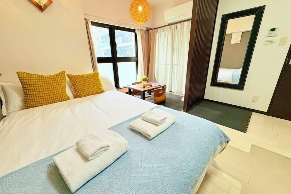A hotel room with a large bed, two pillows, a bedside table with a lamp, a mirror, and a window with curtains.