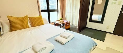 A hotel room with a large bed, two pillows, a bedside table with a lamp, a mirror, and a window with curtains.