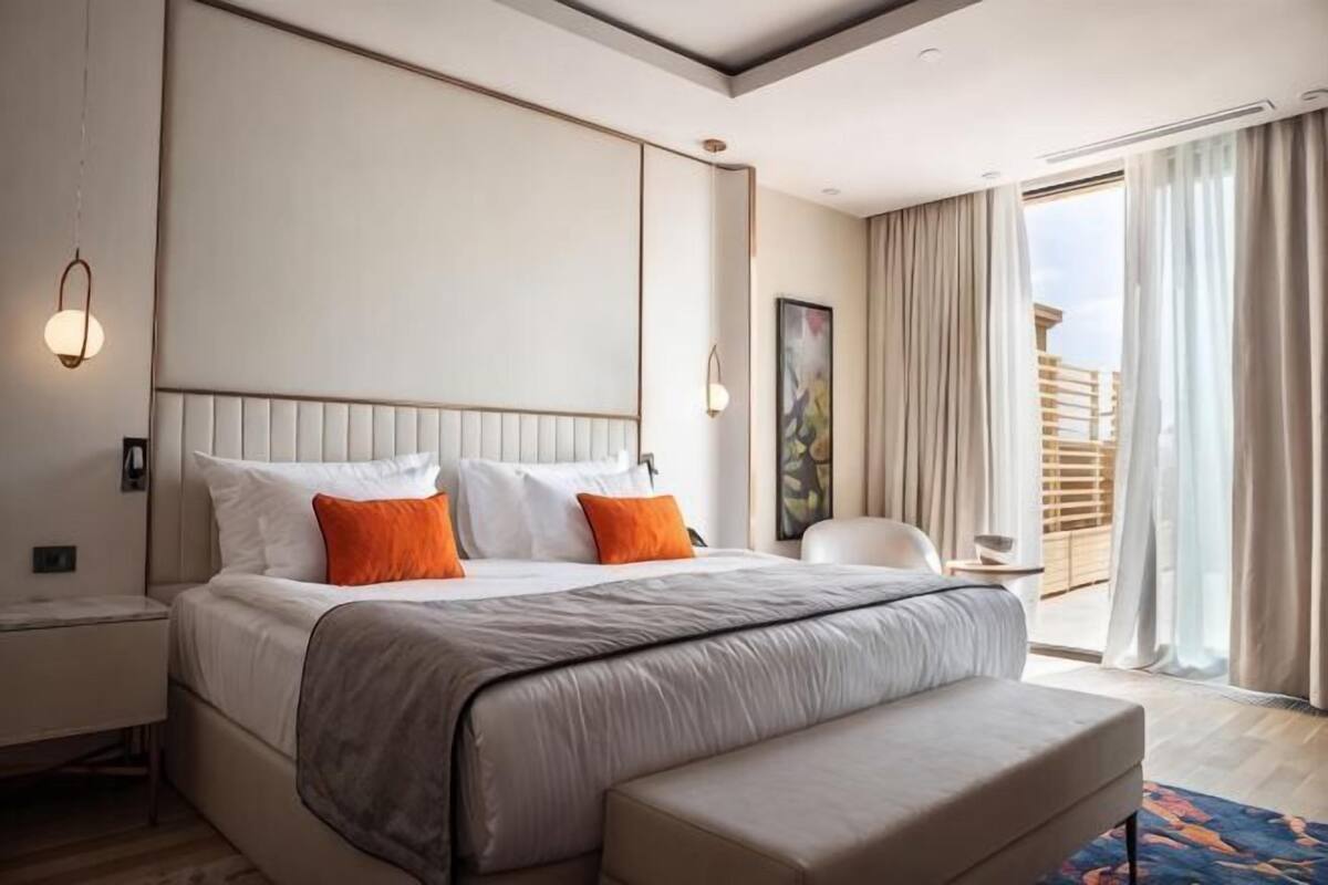 Ein Hotelzimmer mit einem großen Bett, einem Nachttisch, einem Sessel, einem Balkon mit Vorhängen und einem Bild an der Wand.