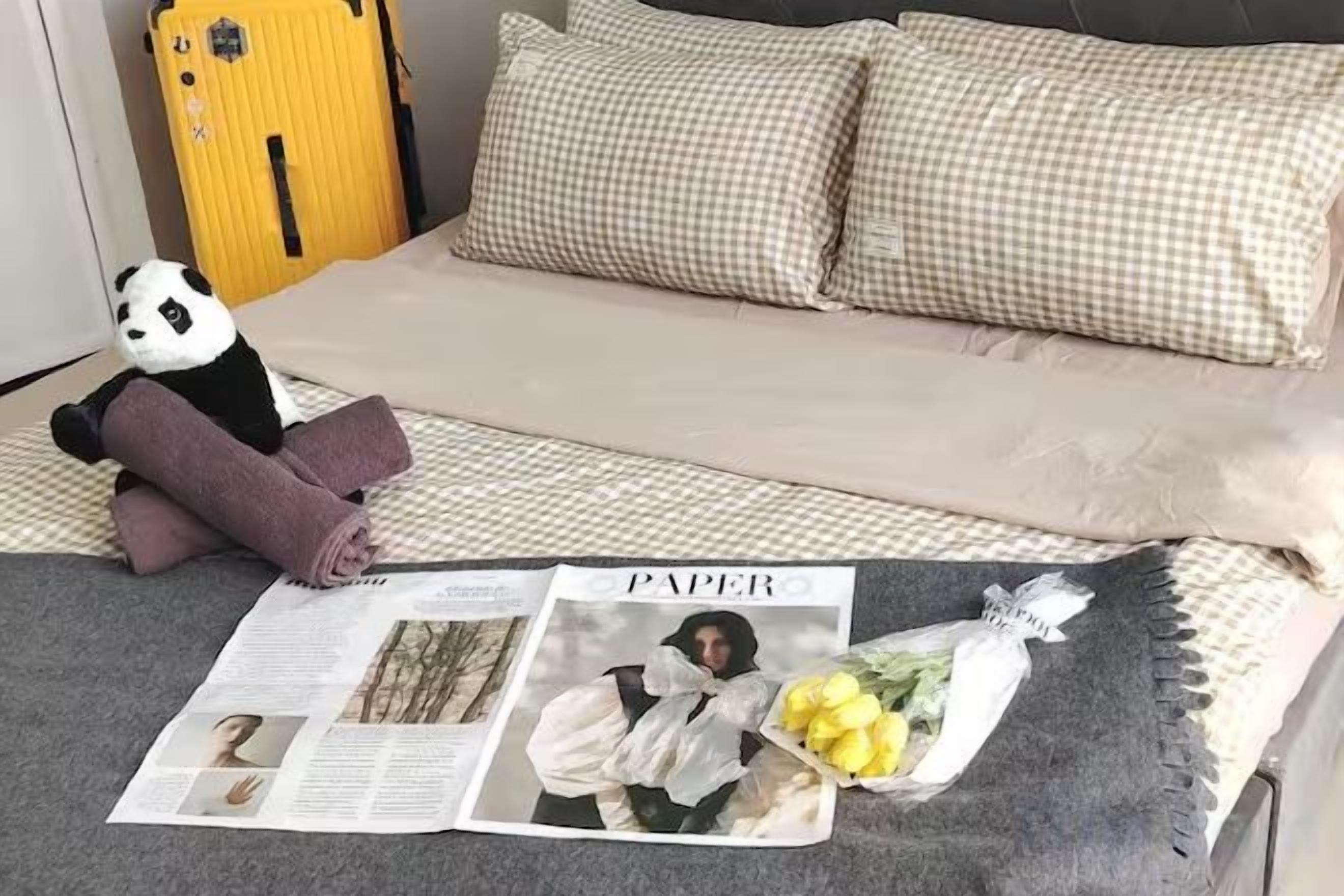 A bed with a checkered pillow, a stuffed panda toy, and a newspaper.
