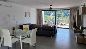 Una sala de estar moderna con una mesa de comedor de vidrio, sillas blancas, un sofá oscuro y un amplio ventanal corredero que da a un espacio exterior.