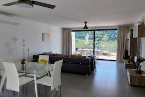 A modern living room with a glass dining table, white chairs, a dark sofa, and a large sliding glass door leading to an outdoor area. - Casa Cidreira (Sao Bartolomeu de Messines)