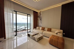 Un salon moderne avec un canapé, un pouf et une table basse, de grandes fenêtres offrant une vue dégagée et un balcon.