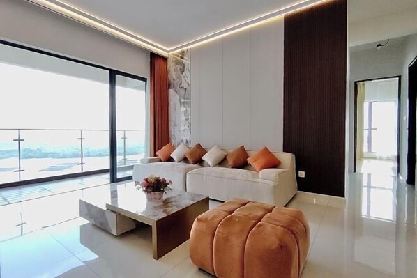 A modern living room with a sofa, ottoman, and coffee table, large windows, and a balcony. - Astonishing Ocean View 4 Bedrooms Huge Balcony 2 Car Park (Johor Bahru)