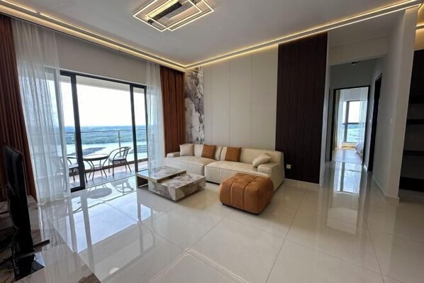A modern living room with a sofa, ottoman, and coffee table, large windows with curtains, and a view of the sea. - Astonishing Ocean View 4 Bedrooms Huge Balcony 2 Car Park (Johor Bahru)