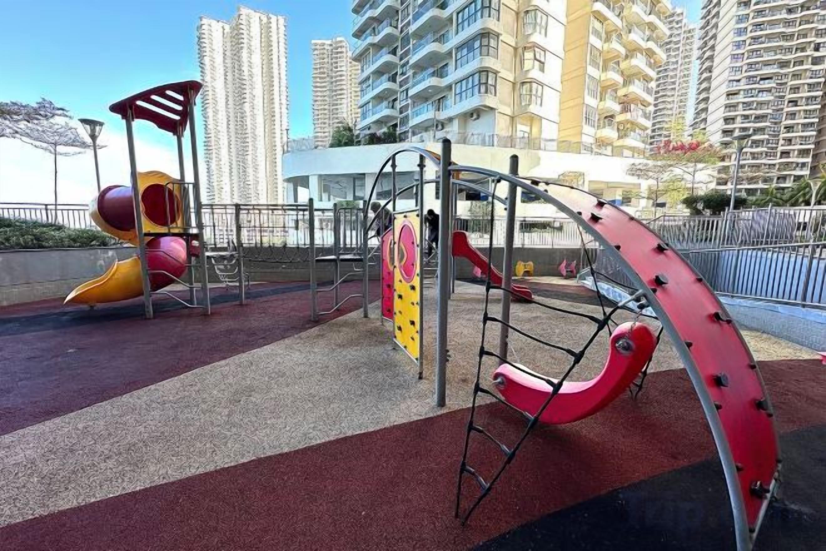 A playground with slides and climbing structures, surrounded by a red safety surface, in an urban setting with tall buildings in the background.