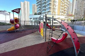Une aire de jeux avec des toboggans et des structures d’escalade, entourée d’une surface de sécurité rouge, située dans un environnement urbain avec de grands immeubles en arrière-plan.